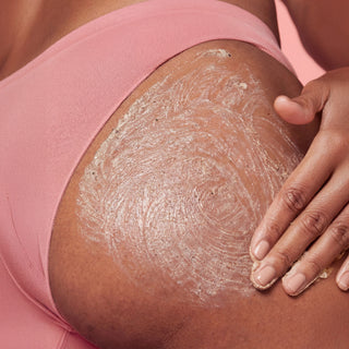 A close-up of a person's hip and upper thigh wearing a pink sports bra, with a white cream or exfoliating scrub being rubbed into the skin by a hand.