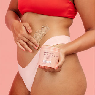 A woman in a red bikini top and pale pink bikini bottom applies a beige scrub to her hip while holding a jar of Express-o Whipped Body Scrub against a solid pink background.