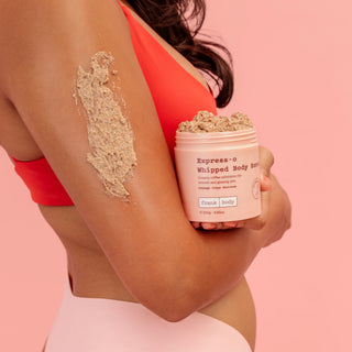 A woman in a coral sports bra holds a jar of Express-o Whipped Body Scrub while showing a thick beige scrub applied to her upper arm against a solid pink background.