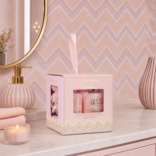 A pink gift box for 'The Luxury Bathing Club' set containing two pink bath products, displayed on a bathroom counter with a lit candle, pink towels, a gold-framed mirror, and a pink striped vase.