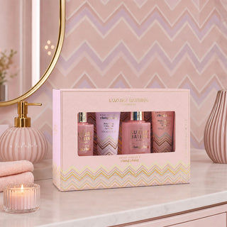 Luxury Bathing London 'Ylang Ylang' bath set in a pink gift box, displayed on a bathroom counter with a pink chevron-tiled wall, gold mirror, and bath accessories.