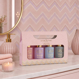 A pink gift box containing four luxury bath products from The Luxury Bathing Company, displayed on a bathroom counter with a mirror, candle, and towels in the background.