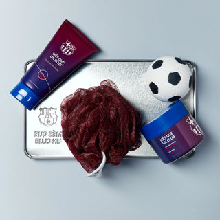 Produse de baie si ingrijire a parului cu tema FC Barcelona, incluzand sampon, ceara de par, bila de baie si burete de baie, asezate pe un capac metalic impreuna cu o minge de fotbal