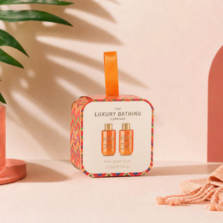 A square gift box for The Luxury Bathing Company Mini Body Duo, featuring a colorful chevron border, an orange ribbon handle, and a white front label showing two orange bottles. The box is styled on a beige surface with soft shadows, palm leaves, and a peach-colored prop.