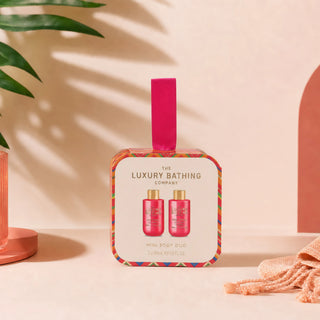 The Luxury Bathing Company Mini Body Duo gift set in a decorative square box with a bright pink ribbon, displayed on a warm beige surface with soft shadows and decorative props.