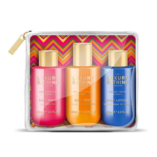 A clear zip pouch containing three Luxury Bathing Company travel body care bottles: a pink body wash bottle, an orange body mist bottle, and a blue body lotion bottle, set against a plain white background.