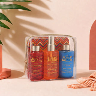 A clear zippered pouch containing three luxury bath products from The Luxury Bathing Company: a red hand & body wash, an orange hand & body wash, and a blue body lotion, displayed on a beige surface with decorative shadows and props.