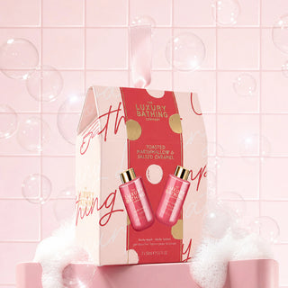 A boxed bath gift set from The Luxury Bathing Company, featuring two pink body wash and body lotion bottles with gold caps displayed on the front of the package. The box is pink with red script and gold foil accents, set against a pink tiled background with floating bubbles and bath foam.