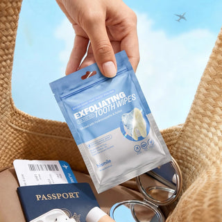 A hand holds a blue and white packet of exfoliating tooth wipes above a woven straw bag, with a passport, sunglasses, and an airplane visible in the background against a blue sky.