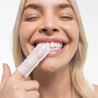 A close-up of a smiling blonde woman with her eyes closed, holding a translucent white finger wax strip against her lower lip with one hand, on a clean white background.